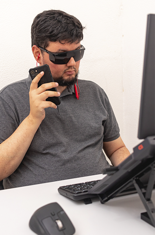 Gustavo Amorim é um homem com deficiência visual. Ele está numa mesa com um notebook à sua frente e segura um celular posicionado próximo ao ouvido.